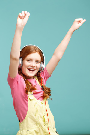 Immaculate song. Joyful little red-haired girl holding her hands up in the air and listening to music while evincing happiness.の写真素材
