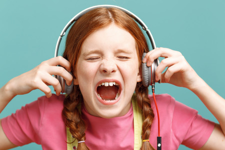 Out loud. Close up of emotional young red-haired girl keeping her headphones and evincing herself while listening to music ob blue background.の写真素材