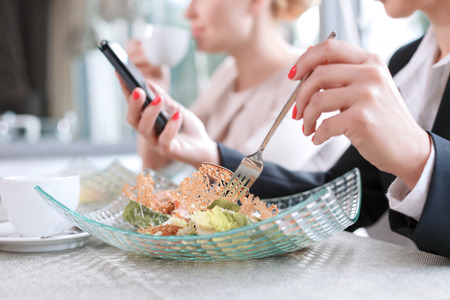 Close up photo of hands on a woman holding a fork eating a salad and a mobile phone reading some important emails, in a restaurant during business lunch selective focusの写真素材