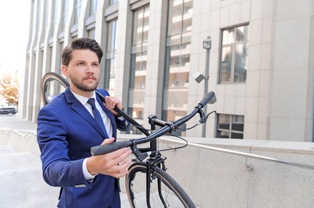 Nice handsome bearded businessman looking up and touching handle of the bicycle while holding it on...