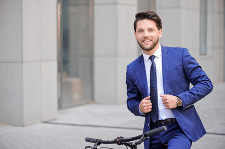 Nice bearded businessman keeping hands on his suit and smiling while riding bicycleの写真素材