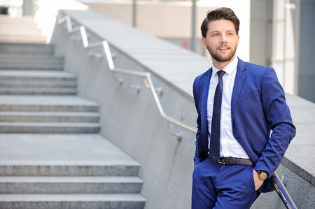 Pleasant bearded businessman putting  his hands in pocket and leaning on the handrail with office center on backgroundの写真素材