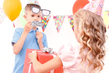 What a surprise. Portrait of little girl giving birthday present to her male friend holding paper party signs.の写真素材