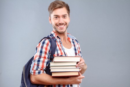 Happy students life. Nice cheerful young student holding books and expressing gladness while standing isolated on gray background.の写真素材