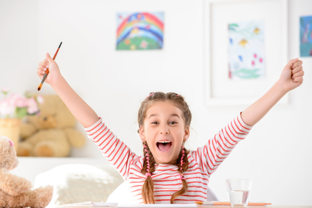 On the edge of joy. Delighted little girl holding her hands up and holding painting brush while feeling completely happyの写真素材