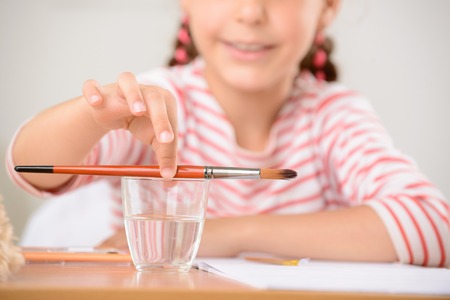 Time to have rest. Pleasant little girl putting brush on glass with water and sitting at the table while expressing positivity.の写真素材
