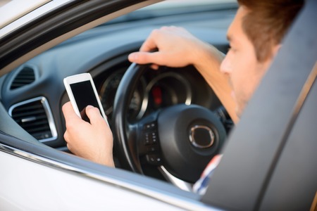 Be modern. Nice upbeat young man holding mobile phone and holding steering wheel while driving his...