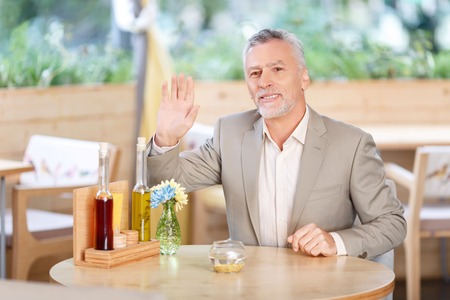 Hello. Pleasant cheerful handsome adult man welcoming you and holding her hand up while sitting in the cafeの写真素材