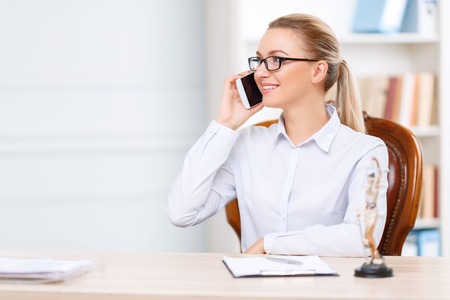 Nice to hear you. Pleasant charming young lawyer sitting at the table and holding mobile phone while having conversation.の写真素材