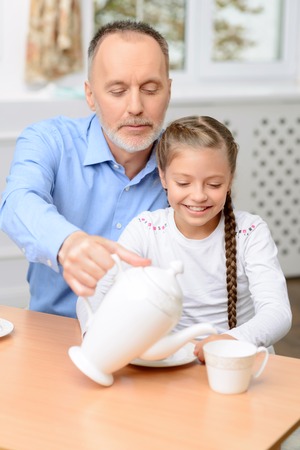 Lets warm our hearts. Pleasant grandfather with little pretty girl sitting at the table and drinking tea while spending time togetherの写真素材