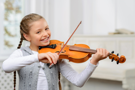 Like doing it. Cheerful blissful pretty little girl holding fiddle bowl and learning to play the violin while smilingの写真素材
