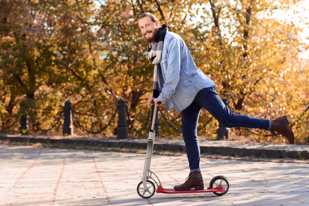 Pleasant handsome blissful bearded man holding the scooter and riding it while feeling delightedの写真素材