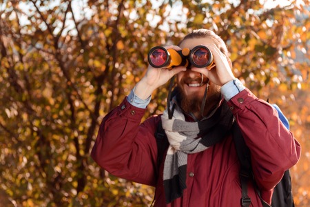 Vivacious smiling bearded tourist holding binoculars and looking through them while travelingの写真素材