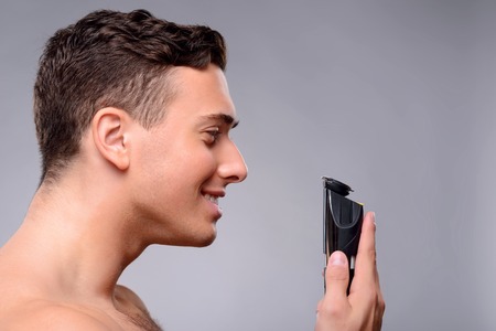 Cheerful positive handsome man holding shaving machine and smiling while looking at itの写真素材