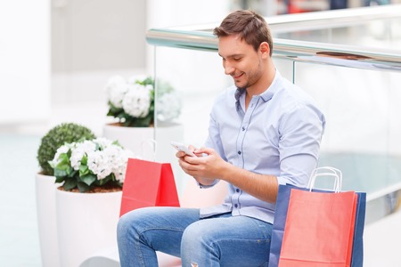 Content smiling agreeable young father holding mobile phone and going to make a call while resting on the benchの写真素材