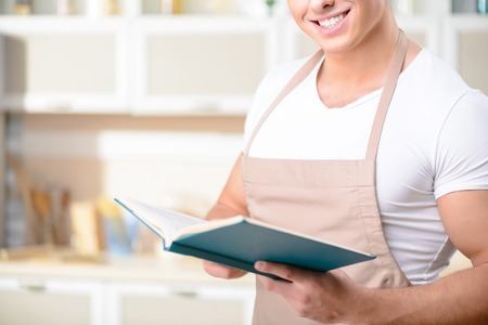 Tough-looking young man with wooden spoon is smiling while checking the recipe book.の写真素材