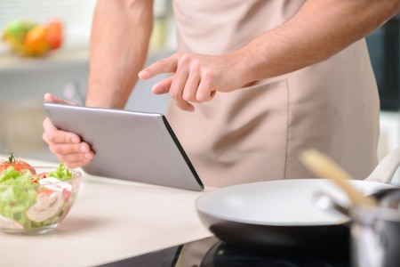 Attractive young strongly built cook is busy with his tablet while standing beside the counter table.の写真素材