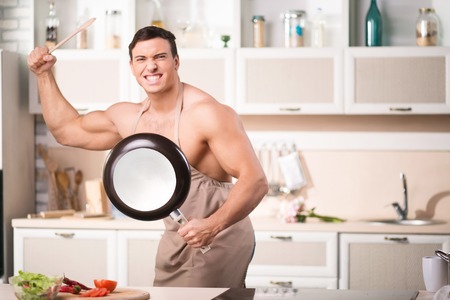 Young attractive strongly built man is posing mockingly with frying pan and wooden spoon.の写真素材
