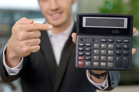 Young attractive salesman is holding a calculator and pointing at incredibly nice price for a carの写真素材