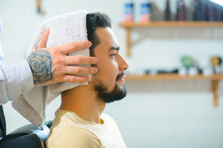 Almost ready. Close up of pleasant content man sitting in the barber shop with professional barber drying his hairの写真素材