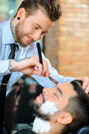 Like my job. Pleasant content smiling barber holding open razor while shaving beard of his clientの写真素材