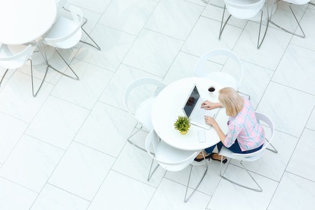 Involved in work. Top view of pleasant blond young woman sitting at the table and using computerの写真素材