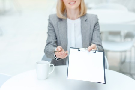 Take it. Selective focus of paper in hands of pleasant professional business woman holding it and giving to you while sitting at the tableの写真素材