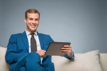 Busy with gadget. Smiling handsome businessman is sitting on the sofa and holding his digital tablet.の写真素材