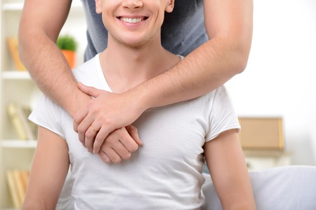 Close up of smiling cheerful guy sitting on the sofa and feeling happy while embracing with his male partnerの写真素材