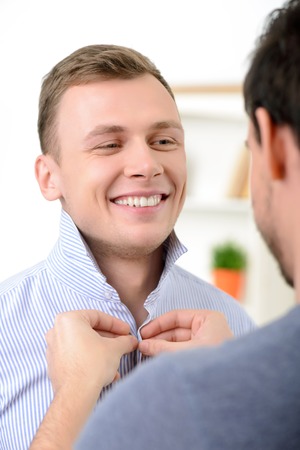 Pleasant delighted guy buttoning the shirt of his male partner while wearing clothesの写真素材