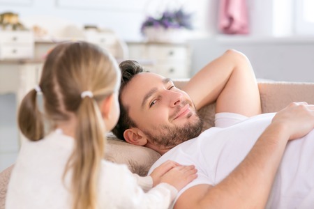 Good morning. Close up of handsome positive father lying on the sofa and going to wake up with his daughter standing nearbyの写真素材