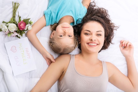 Happy girls. Mom and daughter are smiling while lying in bed and together along with flowers and greeting card.の写真素材