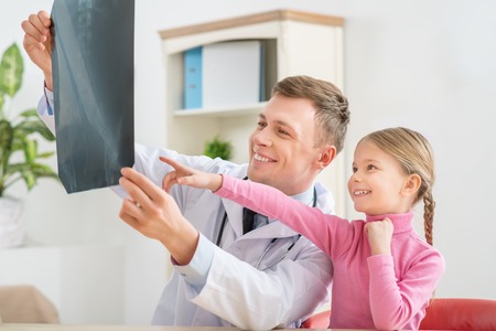 Look here. Cheerful smiling professional pediatrician holding radiogram and sitting on the couch with little girl who is pointing out x-ray photoの写真素材