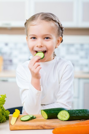Happy childhood. Positive delighted cute little girl eating cucumber and expressing joy while sitting in the kitchenの写真素材
