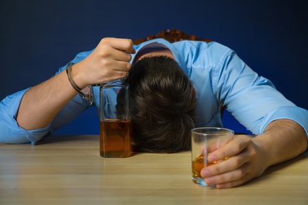 Real addict. Drunk man holding bottle of alcohol and  leaning on the table while sleeping on it