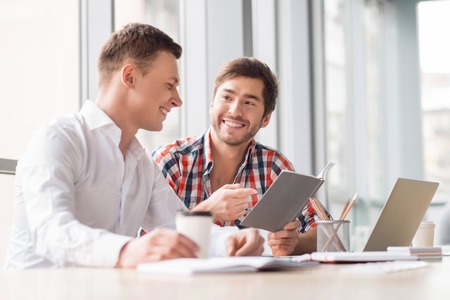 Express your idea. Cheerful handsome men sitting at the table and looking in the notebook while discussing their projectの写真素材