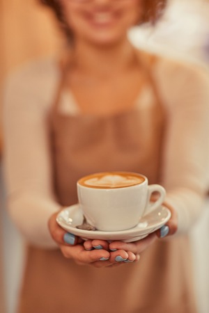 Here you are. Cheerful attractive woman holding cup of coffee and expressing positivity while giving it to youの写真素材