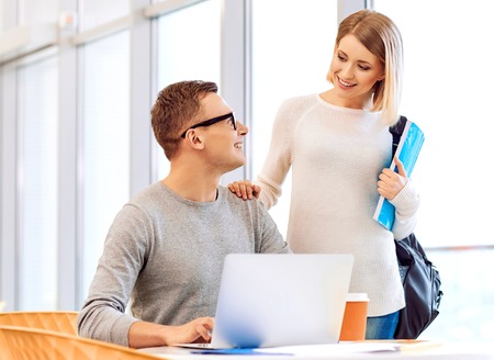 How are you getting on. Pleasant handsome student sitting at the table and working on the laptop while his friend standing nearbyの写真素材