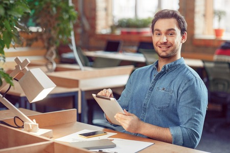 My workplace. Smiling positive content man using tablet and expressing gladness while sitting at the...