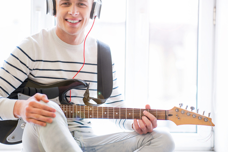 My music world. Cheerful content handsome guy holding the guitar and going to play it while sitting on the window sillの写真素材