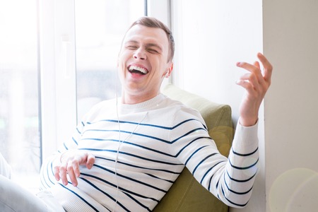 Switch your imagination. Positive emotional guy sitting on the window sill and listening to music while pretending to play the guitarの写真素材