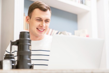 Involved in creative work. Cheerful positive smiling guy  sitting at the table and working on the laptop while expressing joyの写真素材