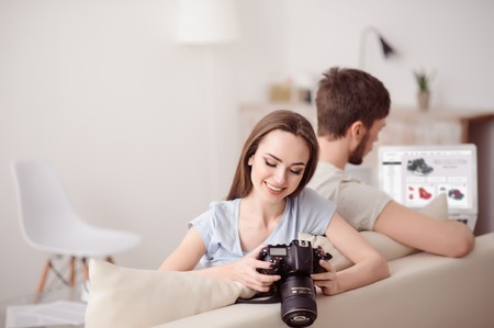 In a high  spirit. Cheerful beautiful   girl  holding photo camera and her boyfriend using laptop shopping online while sitting on the couch togetherの写真素材
