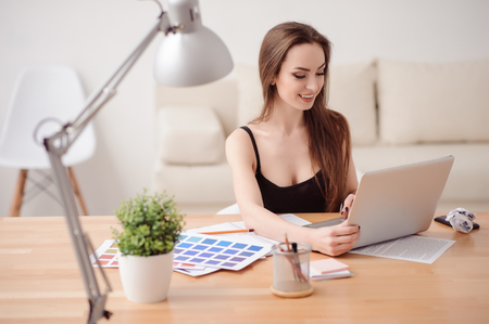 Busy worker. Cheerful beautiful nice girl sitting at the table and using laptop while being involved in workの写真素材