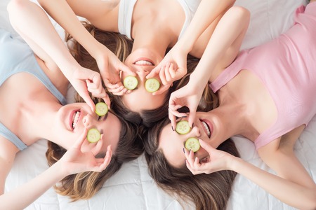 Follow beauty trends.  Positive delighted beautiful girls lying on the bed and holding slices of cucumber on the face while expressing gladnessの写真素材