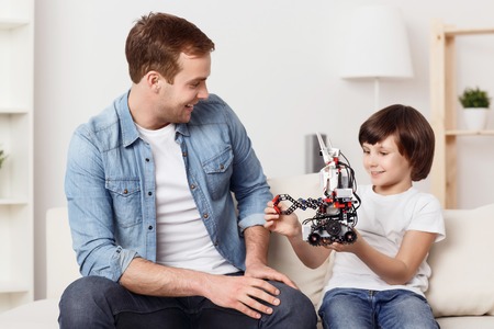 My favorite toy. Cheerful content little boy holding robot and sitting on the couch with his father while smilingの写真素材