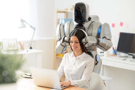 Pleasant pretty smiling woman sitting at the table and working on the laptop while robot wearing headphones on her headの写真素材