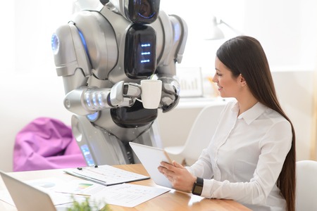 Cheerful nice positive girl sitting at the table and using tablet while robot bringing a cup of...