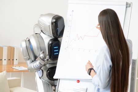 Pleasant girl drawing diagram while modern robot standing nearby and looking at itの写真素材