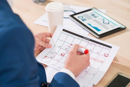 Pleasant man sitting at the table and holding highlighter while marking dates in the calendar